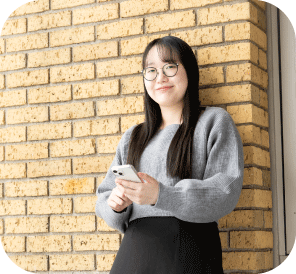 スマホを左手に持ち、壁に寄りかかって立つ 𠮷岡 知美さんの写真